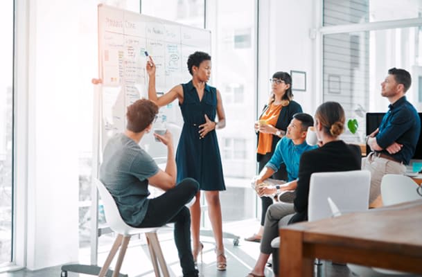 A group of six people are in a modern office, one person is standing and writing on a whiteboard while the others are seated, watching and discussing.