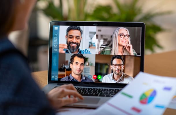 A person holding a document with charts and graphs participates in a video conference call with four individuals displayed on a laptop screen.