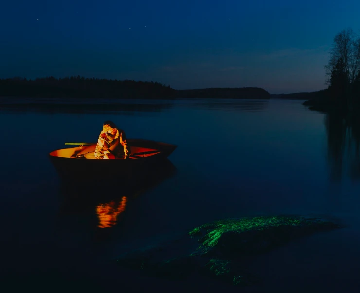 A person is sitting in a small boat on a calm lake at night, illuminated by a light source inside the boat. The surrounding landscape is dark with minimal tree silhouettes in the distance.