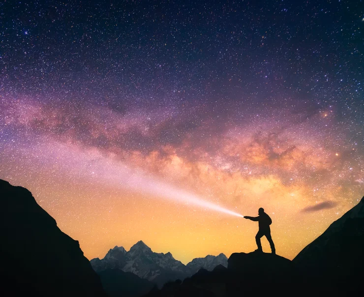 A silhouetted person stands on a mountain peak at night, pointing a flashlight towards the starry sky and the Milky Way. Snow-capped mountains are visible in the background.