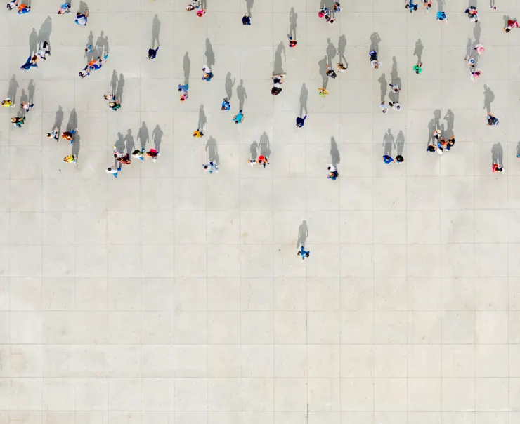 Aerial view of numerous people walking on a light-colored tiled surface, casting distinct shadows.