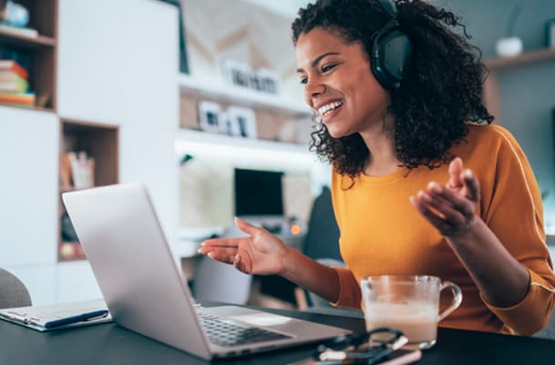 A woman wearing headphones sits at a desk, smiling and gesturing at her laptop with a coffee mug nearby.