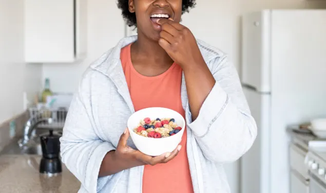 Person standing in a kitchen holding a bowl of cereal with berries, wearing an orange shirt and gray jacket, while eating.