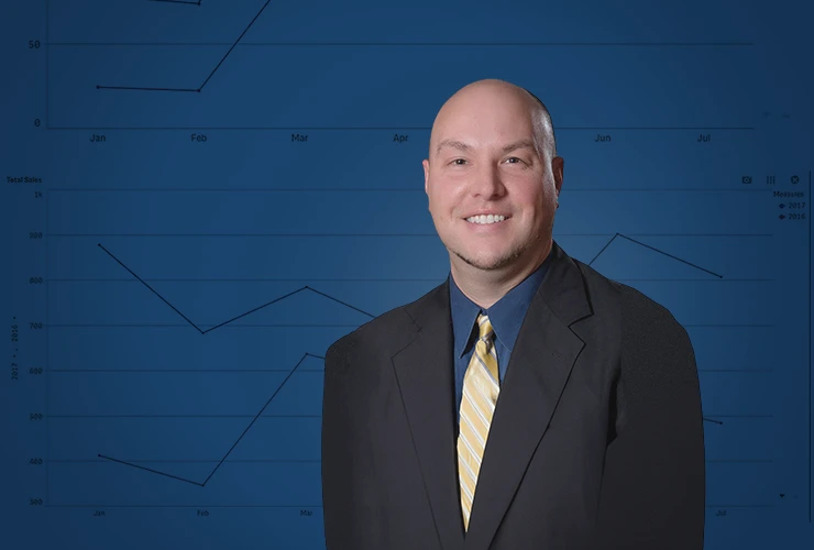 A male in a suit with a tie stands in front of a line graph on a gray background.