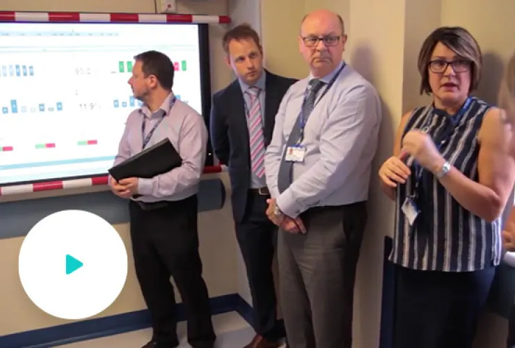 Four people in business attire stand near a screen display. One person points to the screen, another holds a binder, and two others observe.