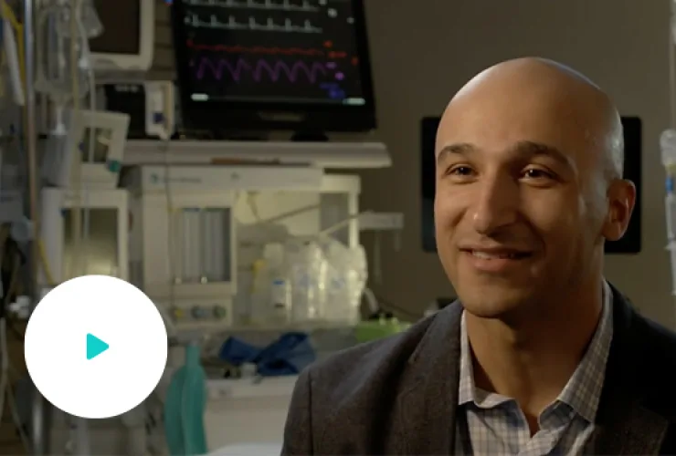 A man in a suit sits in a room with medical equipment, including a monitor displaying a heart rate graph, in the background.