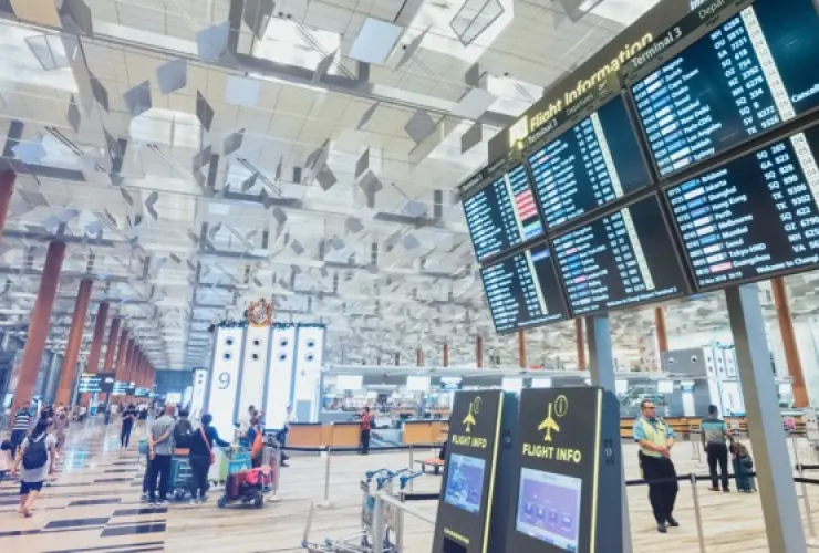 A busy airport terminal with passengers and staff, featuring large digital screens displaying flight information. Several people are pulling luggage or waiting near the check-in counters.