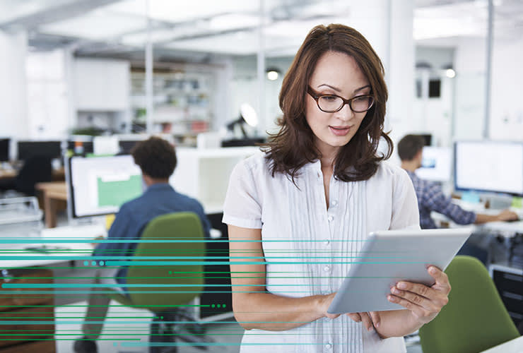 A woman in an office reads from a tablet. Two other people work on computers in the background.