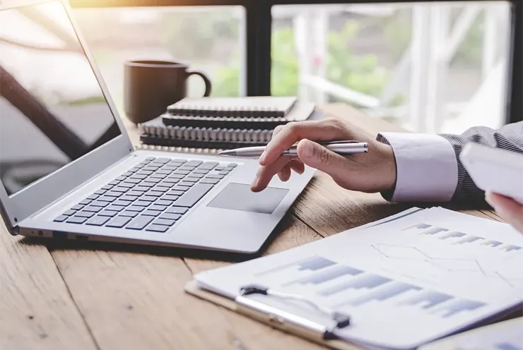 A person uses a laptop and holds a smartphone at a desk with notebooks, a clipboard with charts, and a cup in the background.