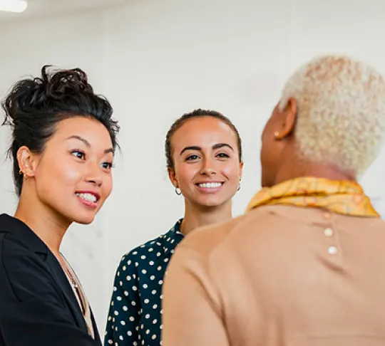 Three people are standing and talking indoors. One person with short blonde hair faces away from the camera while two others, one with a bun and the other with a polka-dot shirt, smile and listen.