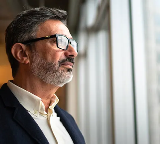 A man with glasses and a beard in a suit jacket looks out of a window with a contemplative expression.