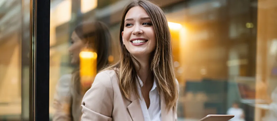 A woman in business attire smiles while holding a tablet, standing outside a glass-fronted building.