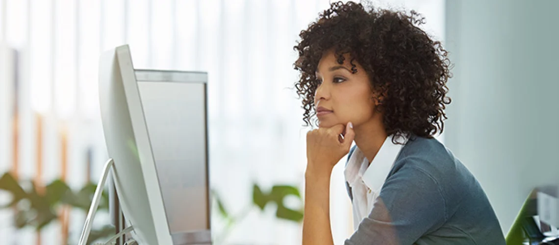 A person with curly hair and a contemplative expression sits at a desk, looking at a computer monitor.