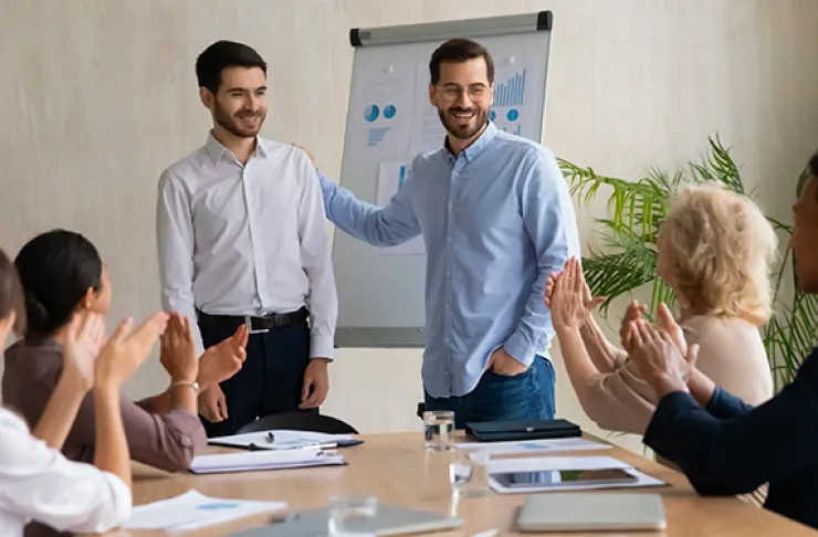 Two men stand near a flip chart as one of them gestures to the other, while colleagues seated around the table applaud.