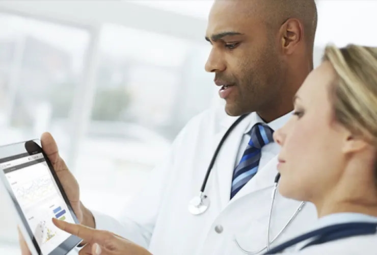 Two doctors, one male and one female, review medical data on a tablet in a bright, modern setting. Both are wearing white lab coats and stethoscopes.