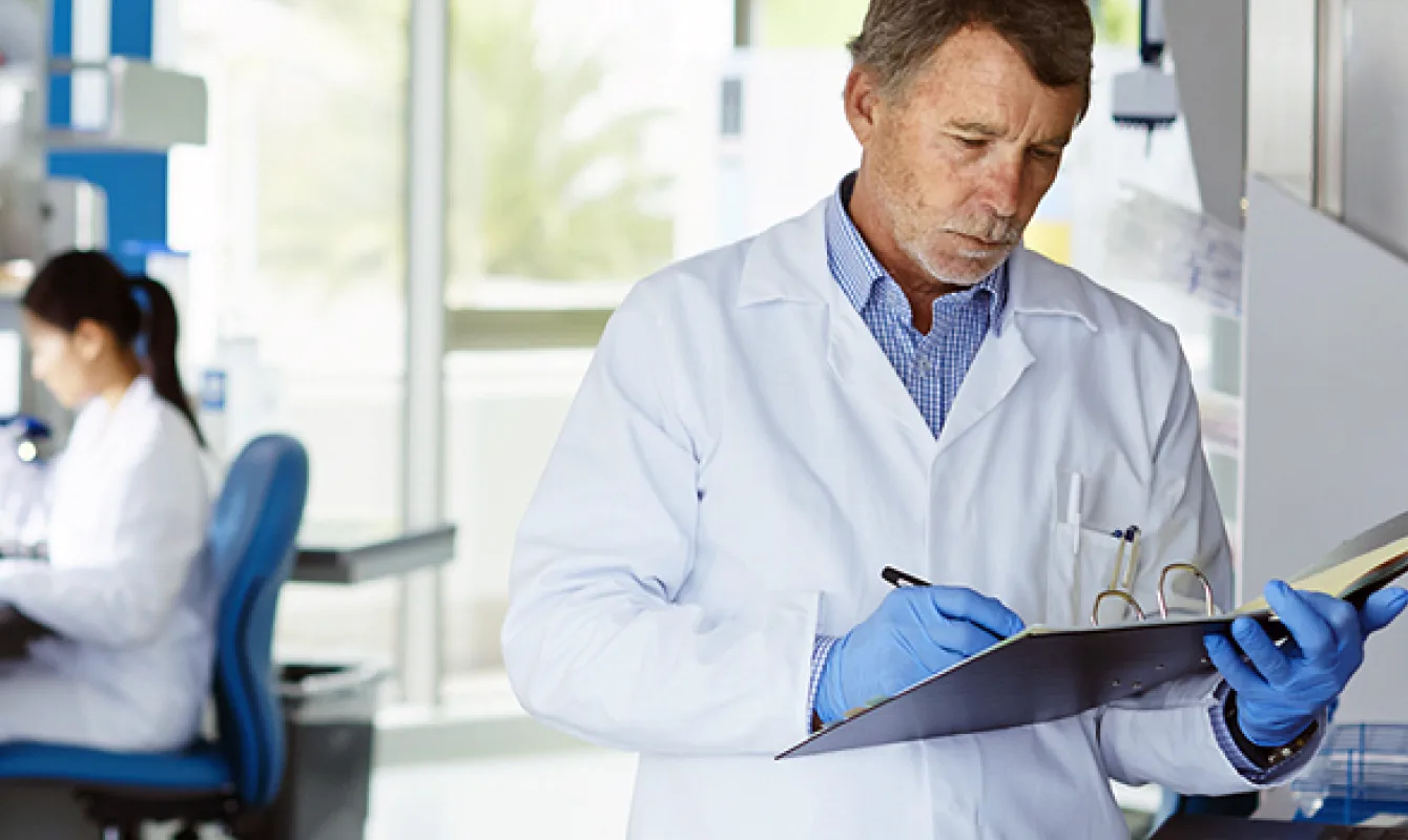 A male scientist in a white lab coat and blue gloves writes on a clipboard in a laboratory, with another scientist working in the background.