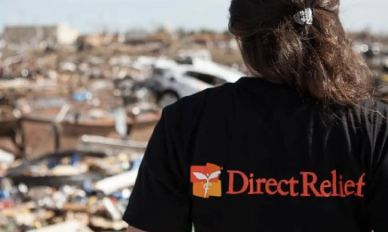 Person wearing a black "Direct Relief" shirt stands facing debris and destruction in a disaster area.