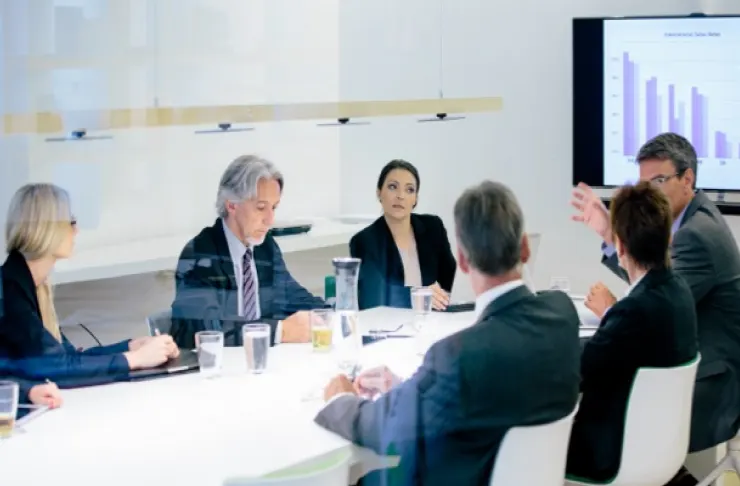 Six business professionals sitting around a table in a conference room, with one person gesturing towards a presentation screen displaying bar graphs.