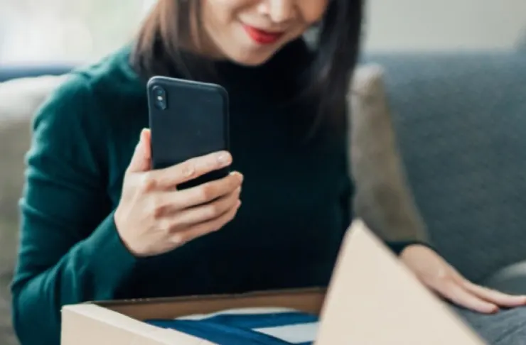 A woman in a green sweater uses her smartphone while looking into an open cardboard box.