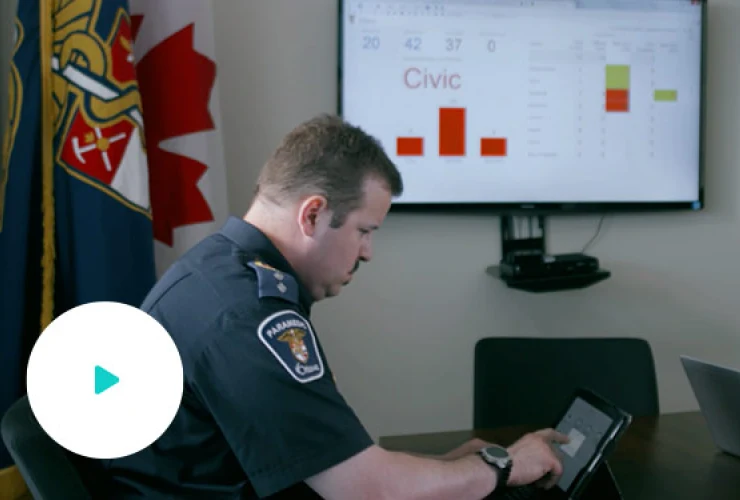 A person in a uniform uses a tablet at a table with a screen displaying charts and the word "Civic" in the background. Flags and official insignia are visible in the room.