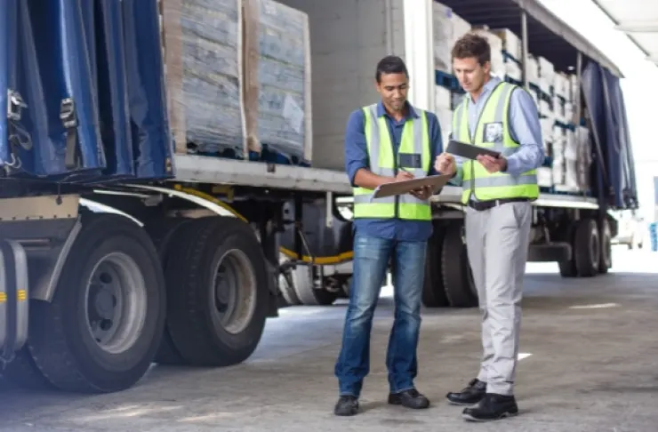 Two workers in safety vests stand next to a truck with an open trailer, examining clipboards.