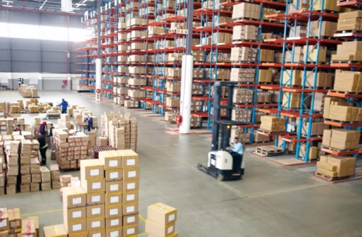 A warehouse with tall shelves stacked with boxes, a few workers sorting packages, and a forklift moving items on the floor.