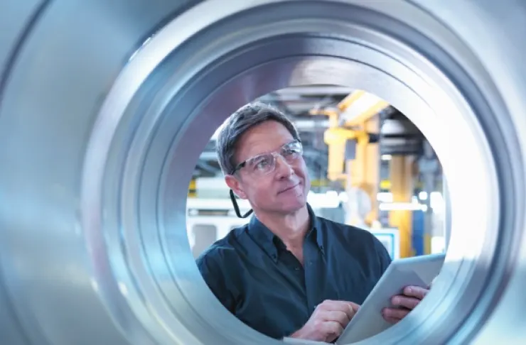 A man wearing safety glasses and holding a tablet stands inside a metal manufacturing facility, looking up through the center of a large metal coil.