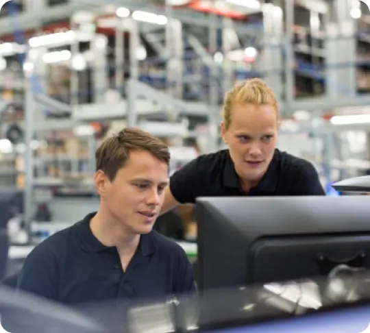 Two individuals in a factory setting, both wearing black shirts, are working together at a computer workstation. The background is filled with industrial equipment and shelving.