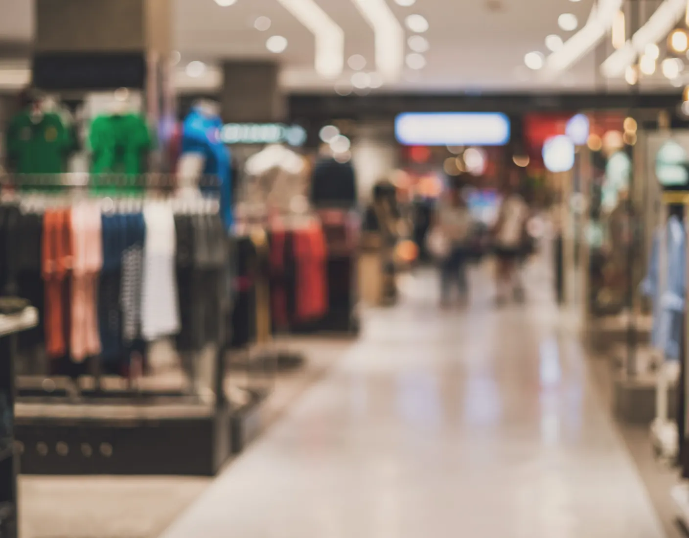 Out-of-focus image of a clothing store interior with racks of garments on the sides and shoppers in the background.