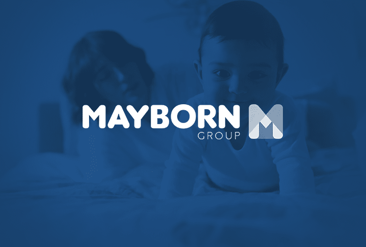 A baby crawling on a bed with an adult in the background. The Mayborn Group logo is displayed prominently over the image.