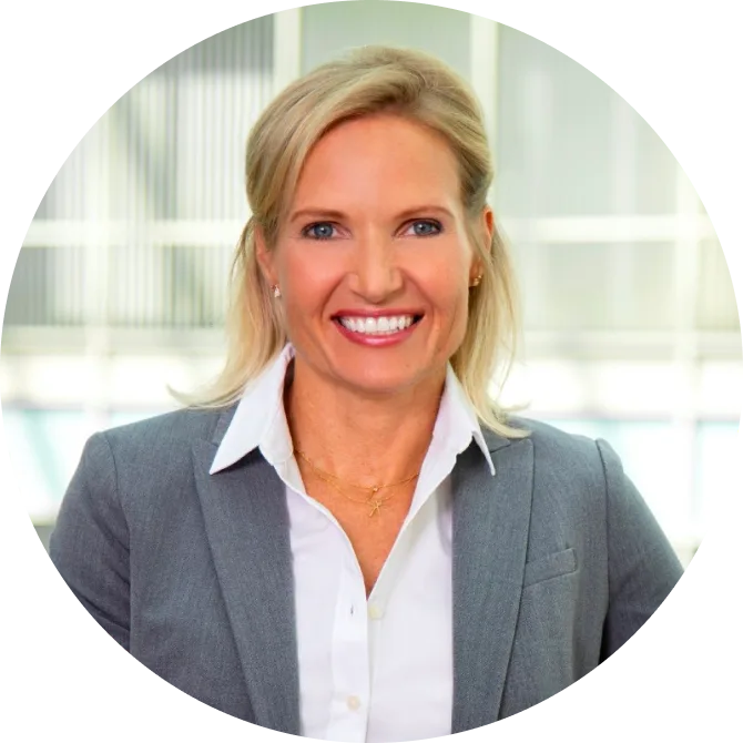 Headshot of Julie Kae, with blonde hair and a friendly smile wearing a white blouse and gray blazer, standing in front of a blurred window in the background.