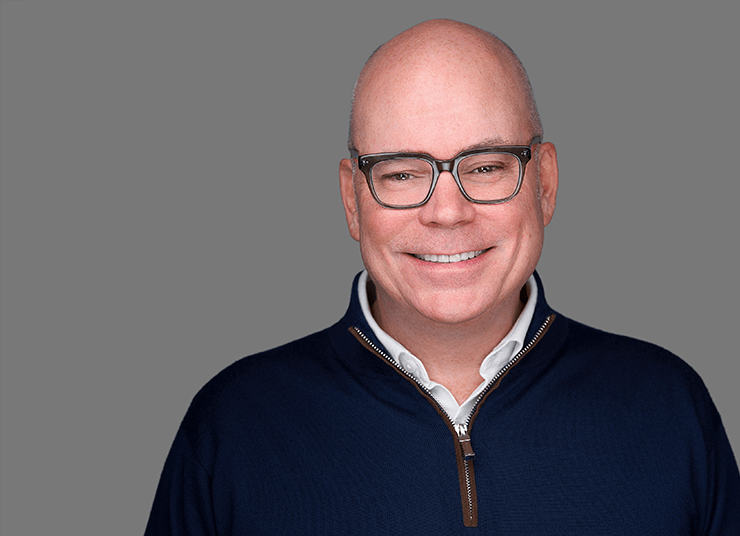 Chris Powell, Chief Marketing Officer at Qlik dons glasses and is wearing a navy sweater over a white shirt, smiling, against a grey background.