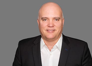 Mike Potter, Chief Technology Officer at Qlik in a dark suit jacket and white shirt stands against a plain gray background, smiling into the camera.