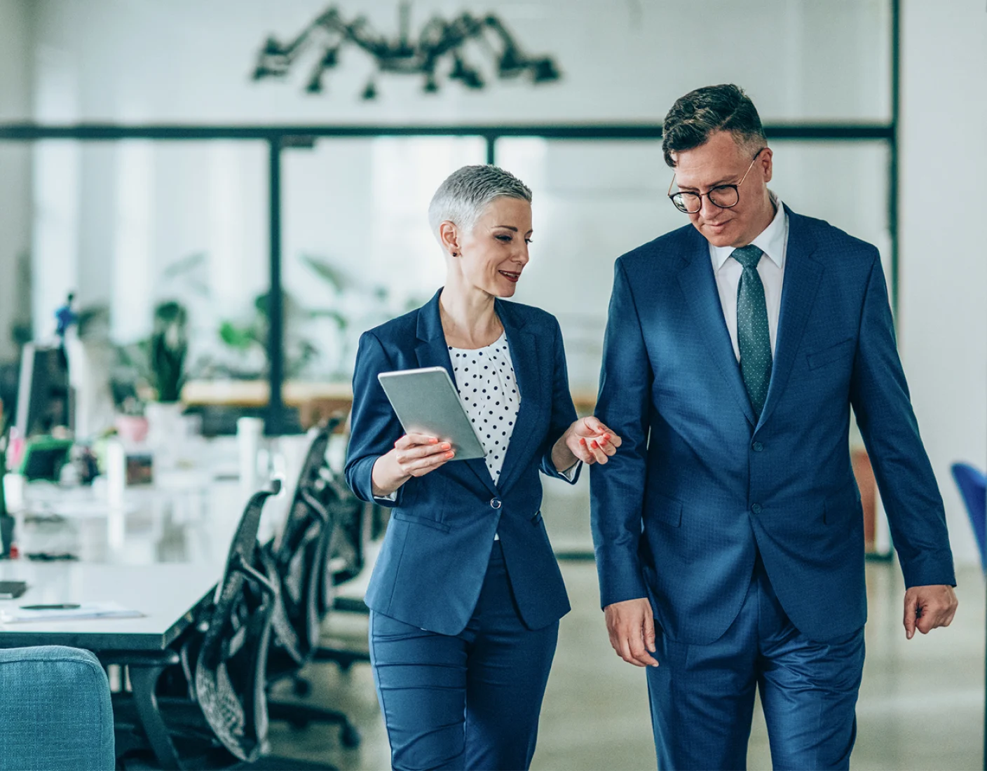 Two business professionals in formal attire walk through an office. The woman holds a tablet and they appear to be discussing something as they walk.