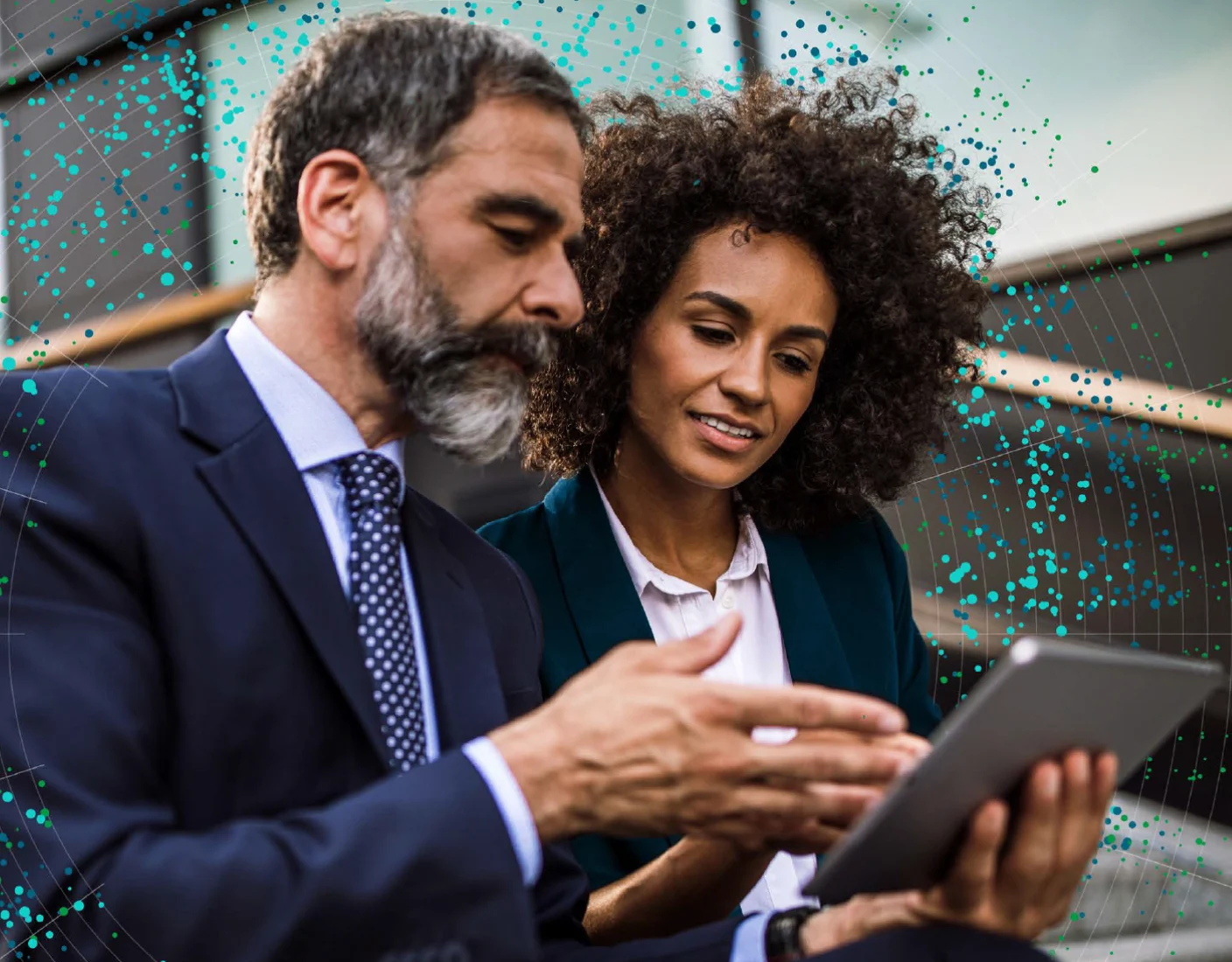 Two professionals, a man with a beard in a suit and a woman with curly hair in business attire, review information on a tablet in an outdoor setting.