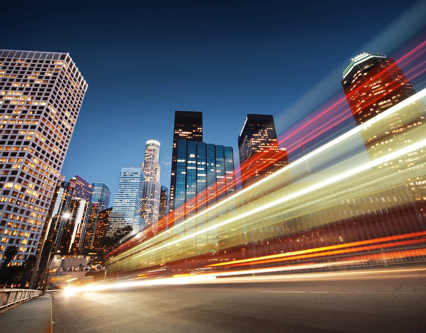 A cityscape at twilight with tall buildings and a long-exposure effect creating streaks of light from moving vehicles in the foreground.