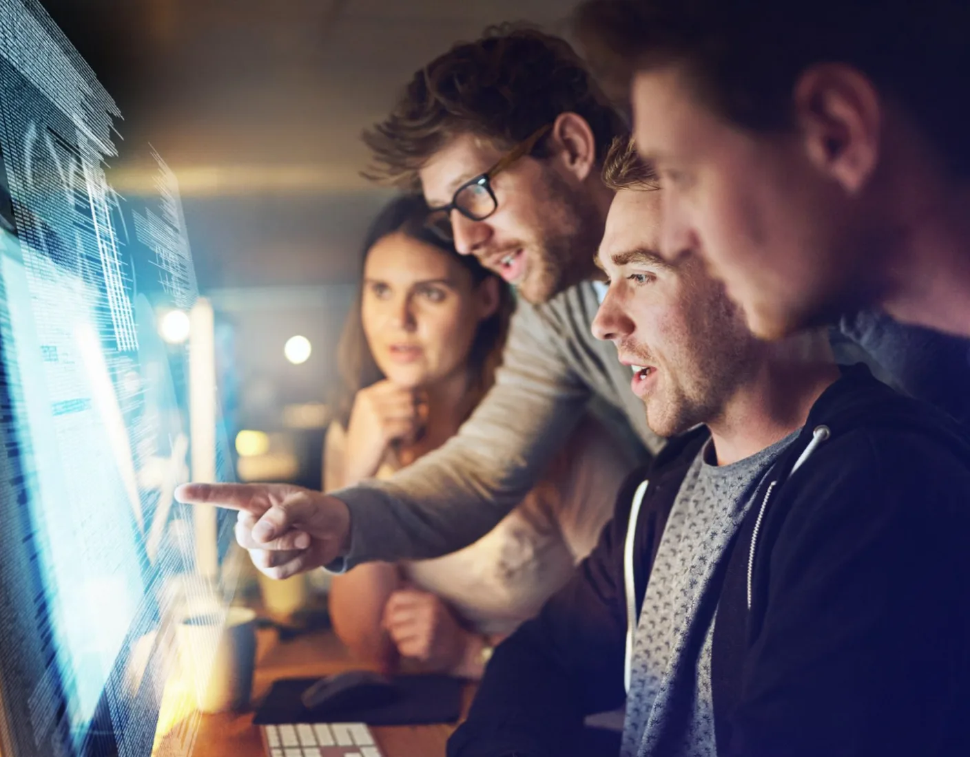 A group of four people look intently at a computer screen, with one person pointing at the display. They appear to be working together on a project.