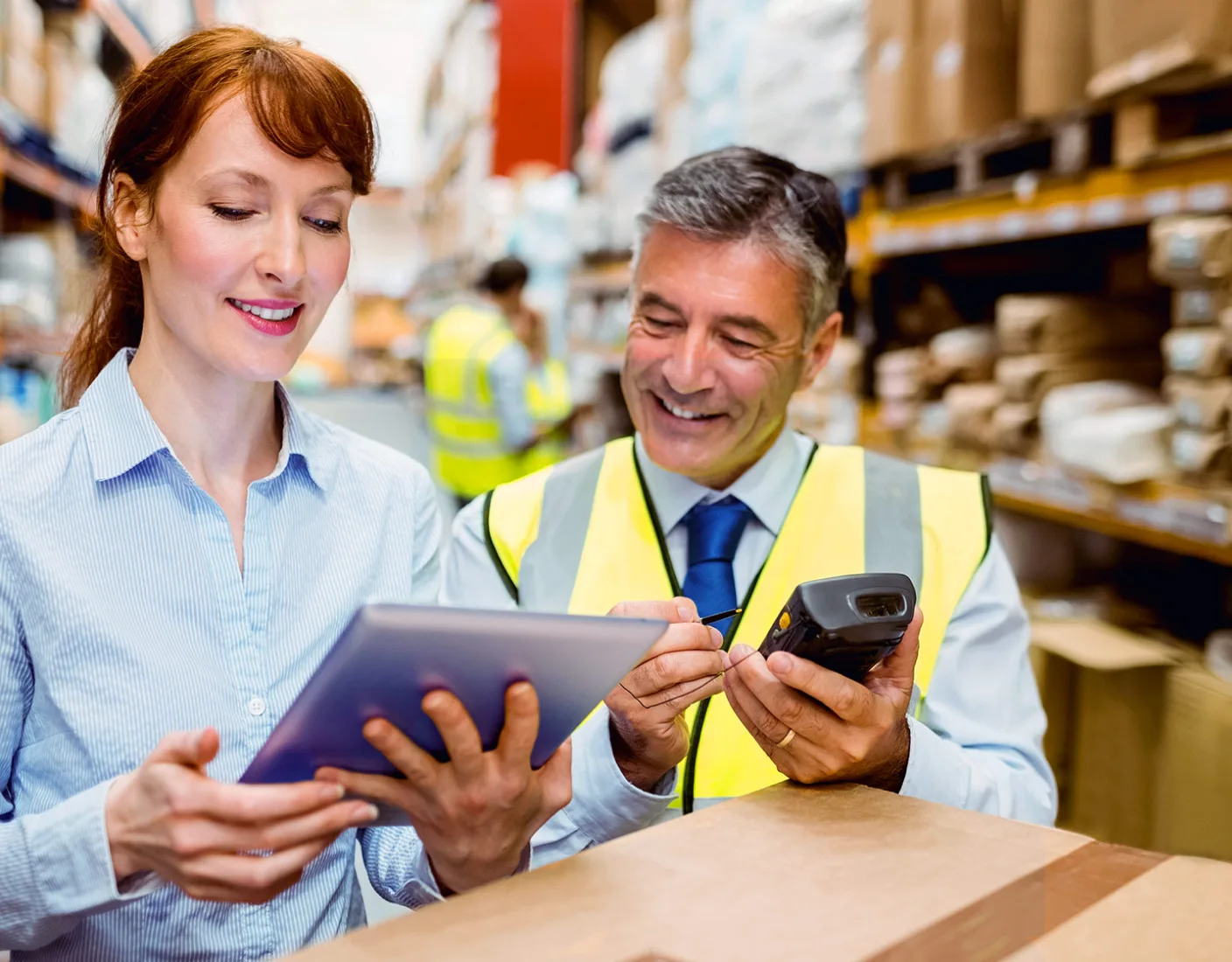 Two warehouse workers, one holding a tablet and the other using a handheld scanner, are smiling while working in a storage area with shelves filled with boxes.