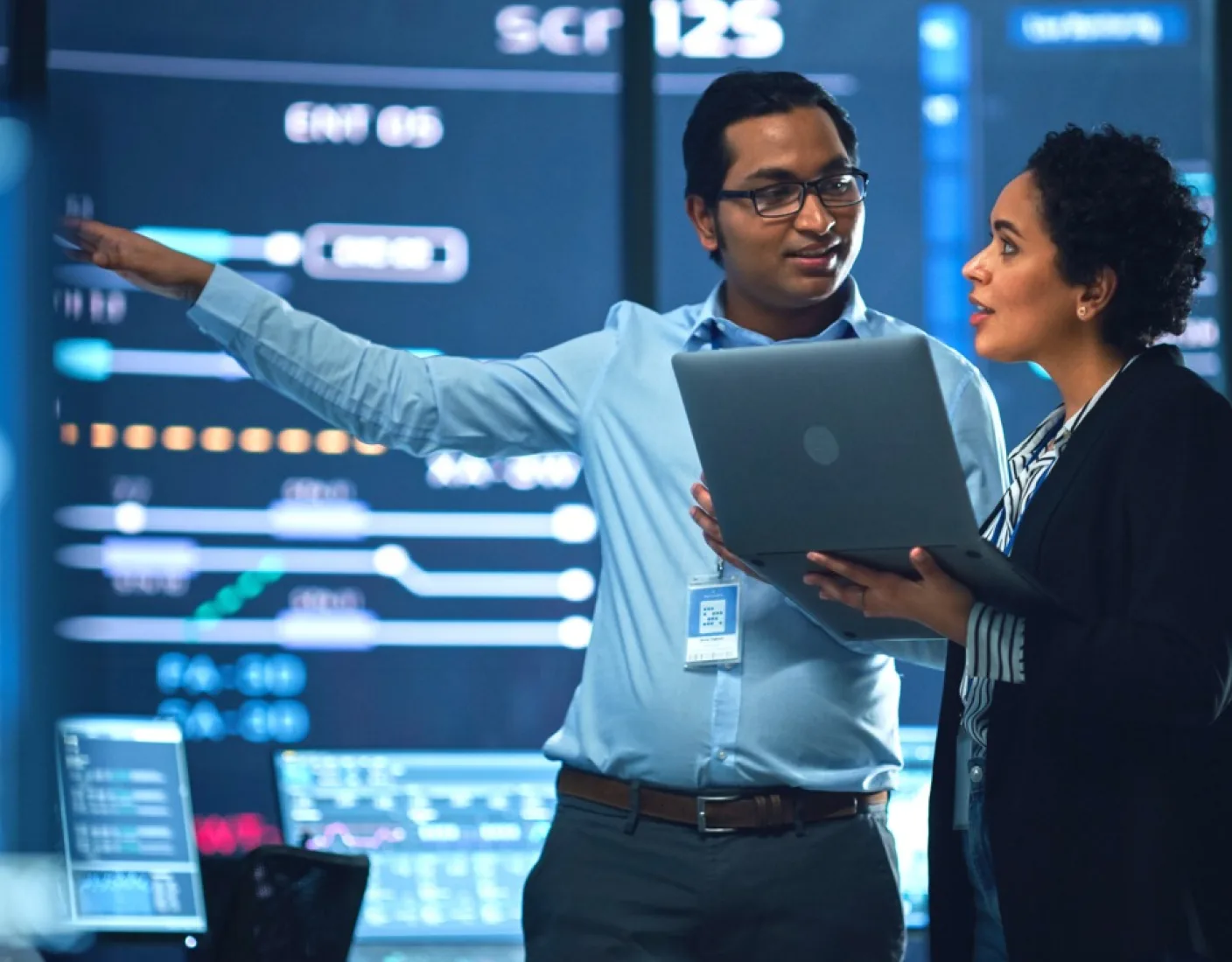 A male and female have a conversation while viewing a large wall-screen displaying stats and charts