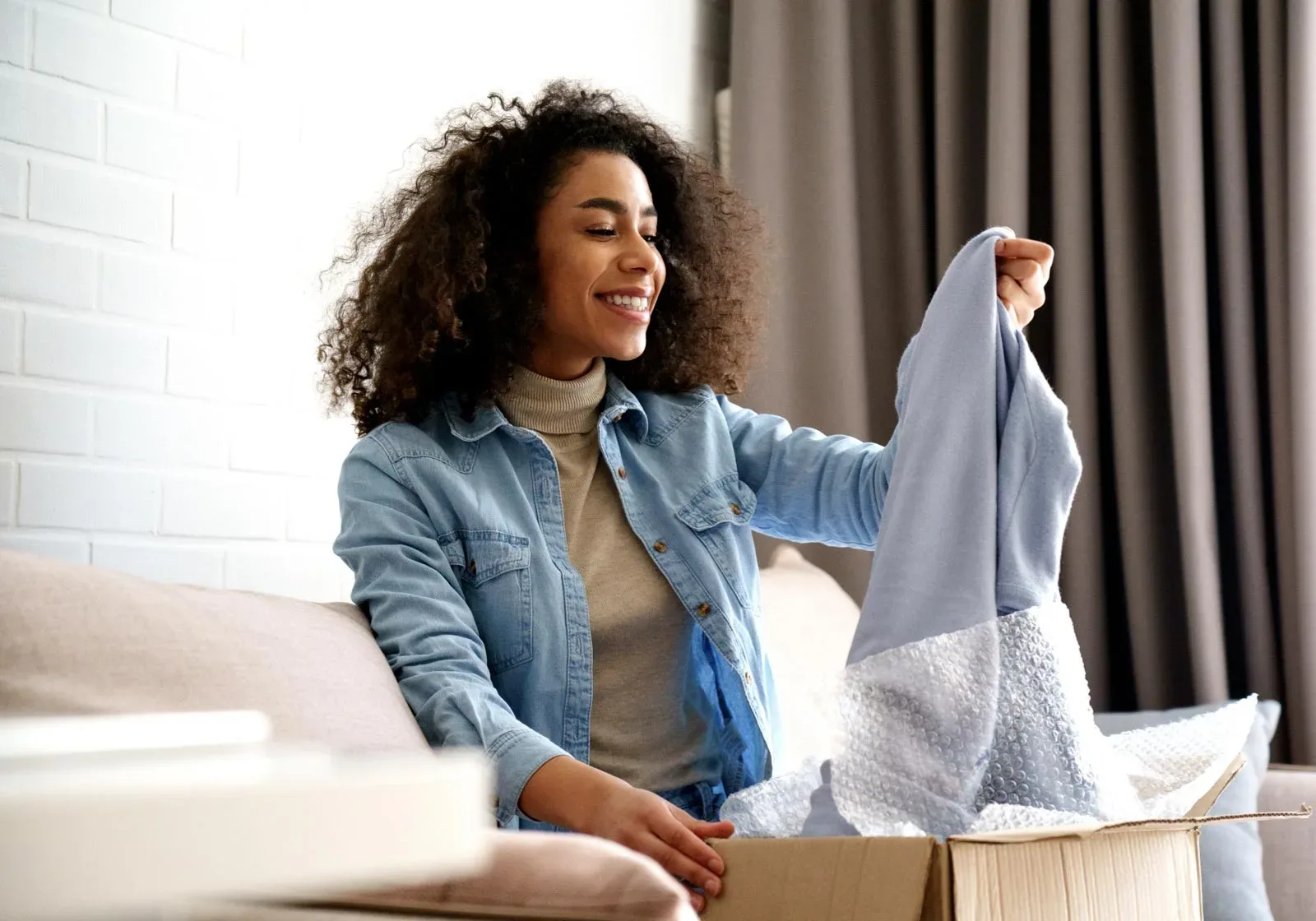 A person with curly hair smiles while unpacking a light blue garment from a cardboard box on a couch.