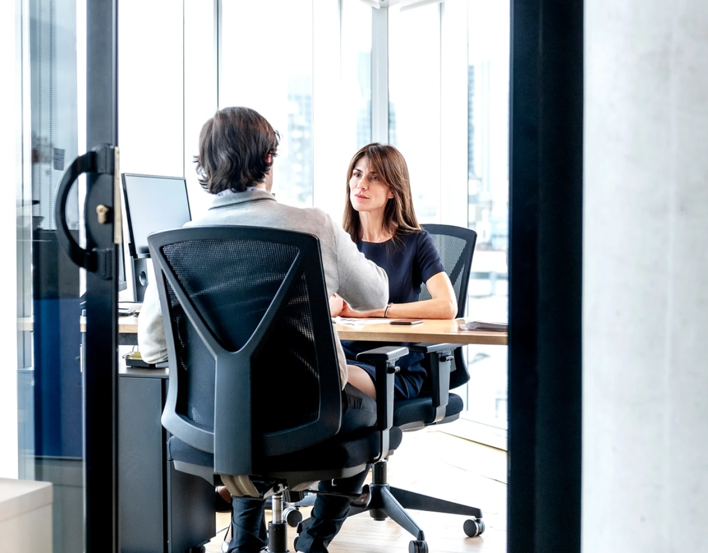 Two people sitting across from each other at a desk in an office. The woman is facing the man and appears to be speaking. Both are seated on modern office chairs.