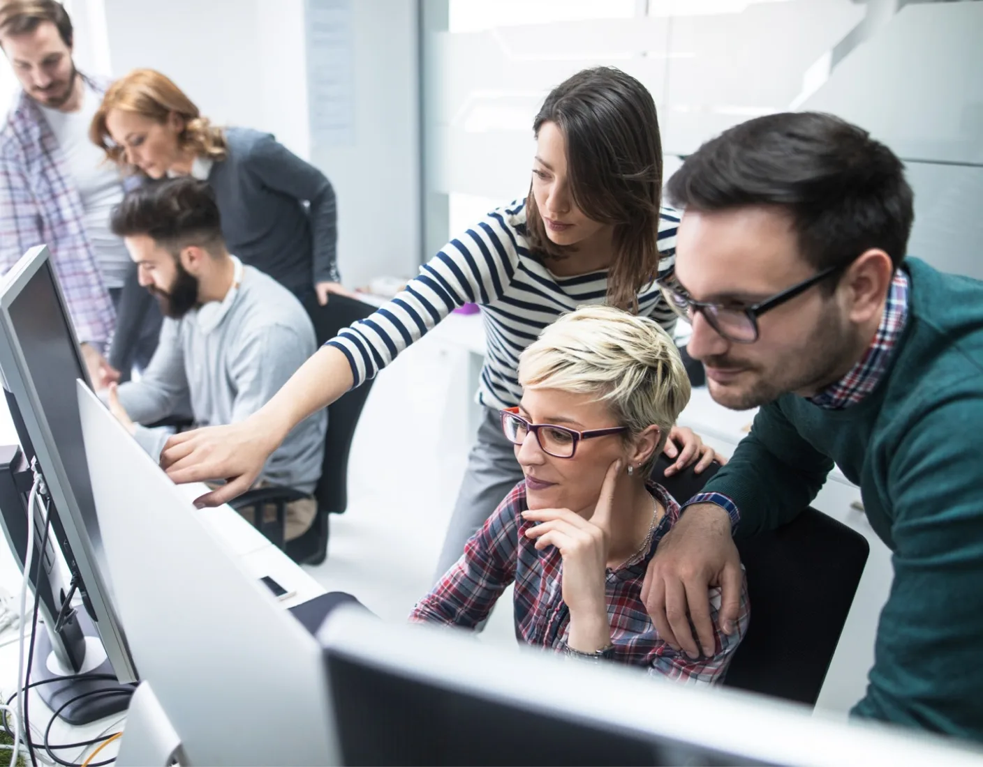 A group of people collaborating around computer monitors in an office setting. One person points at a screen while others look on attentively.