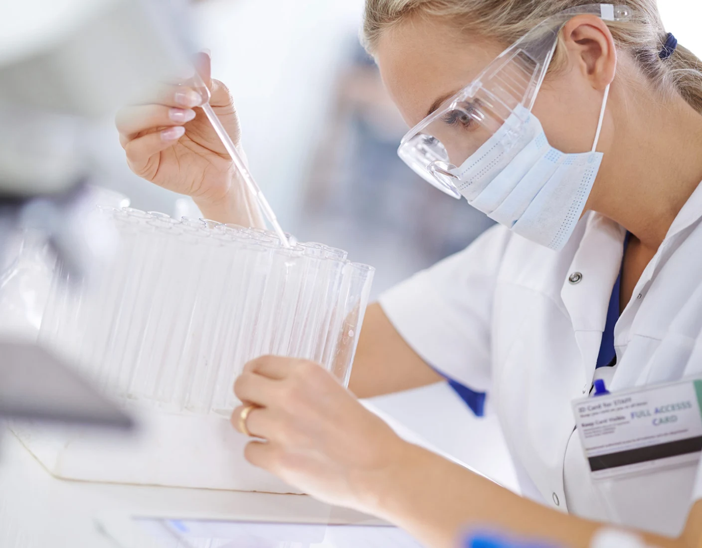 A lab technician wearing safety glasses and a mask uses a pipette in a laboratory setting, surrounded by test tubes.