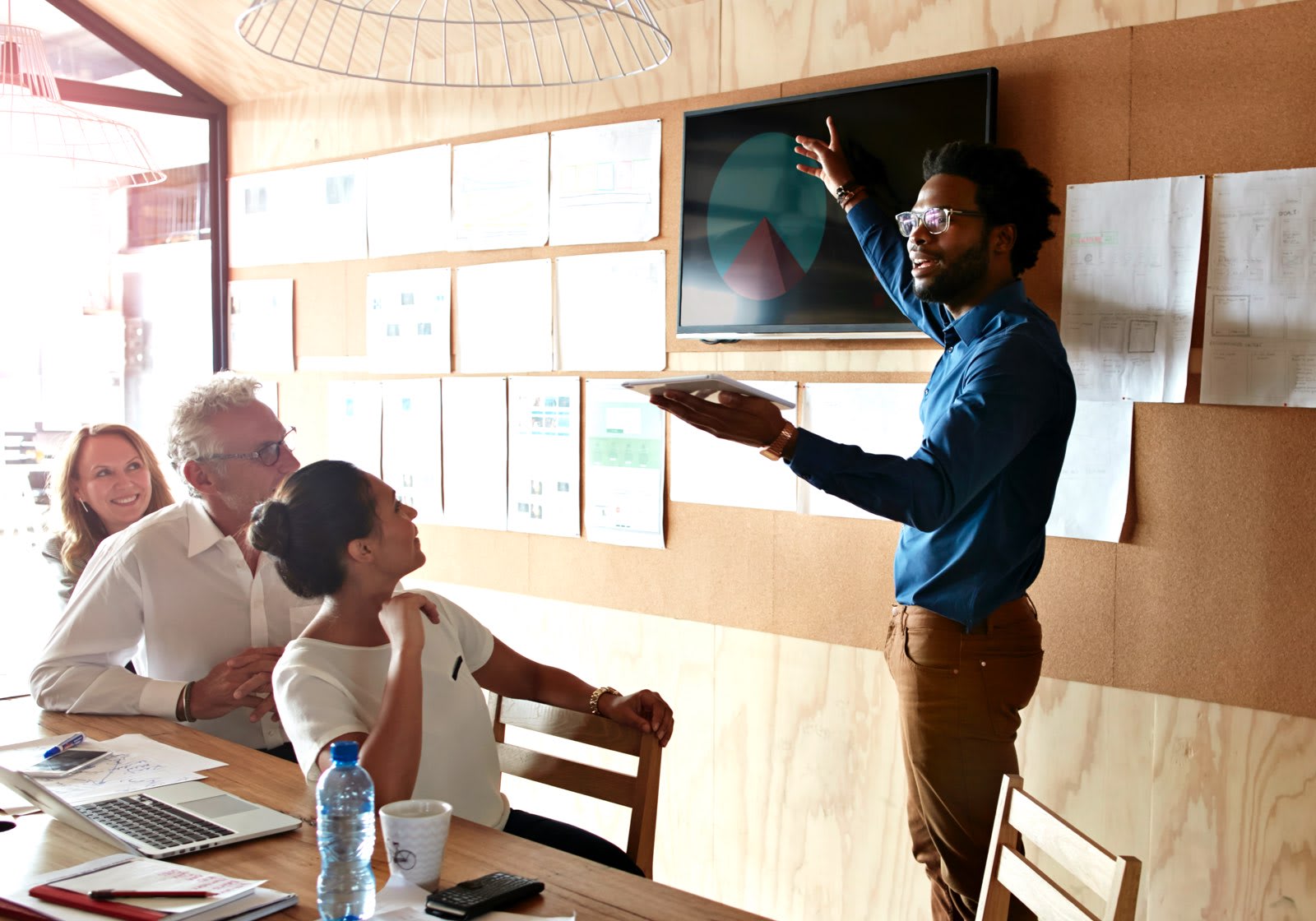 A man stands at the front of a meeting room, gesturing toward a screen displaying a pie chart, while three seated individuals listen attentively.