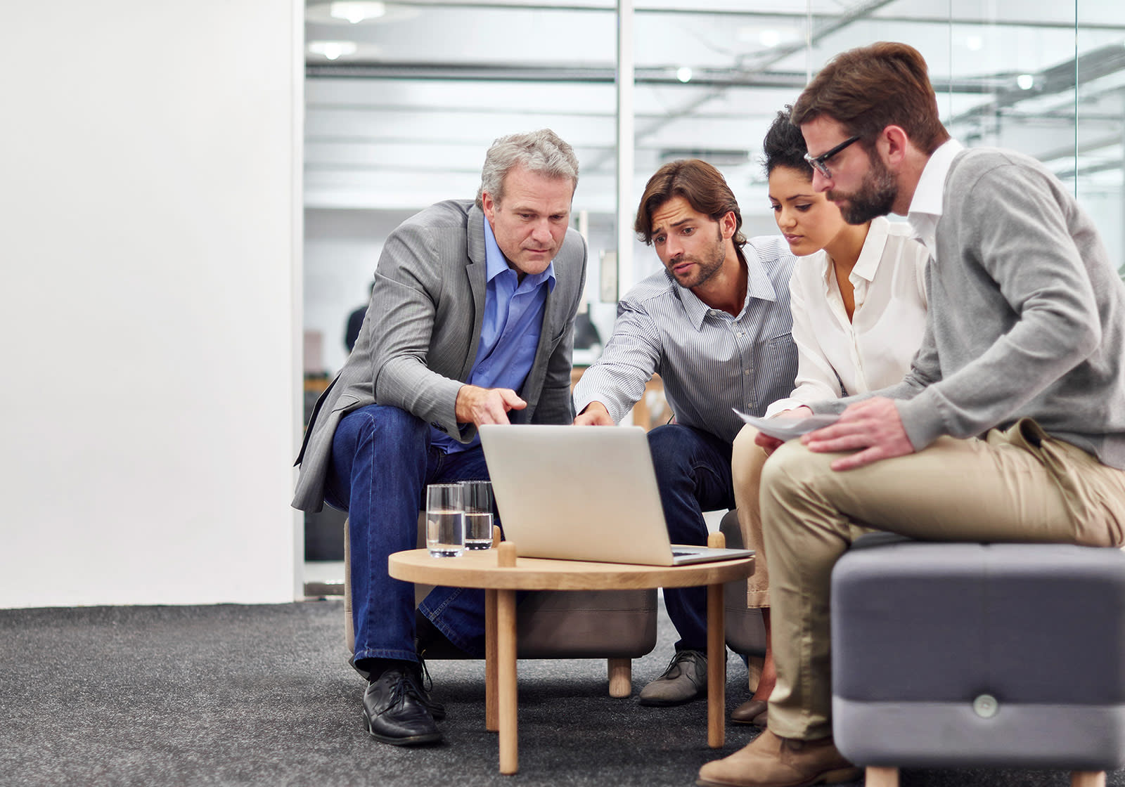 A group of four people sit around a coffee table in an office, discussing and pointing at a laptop screen.