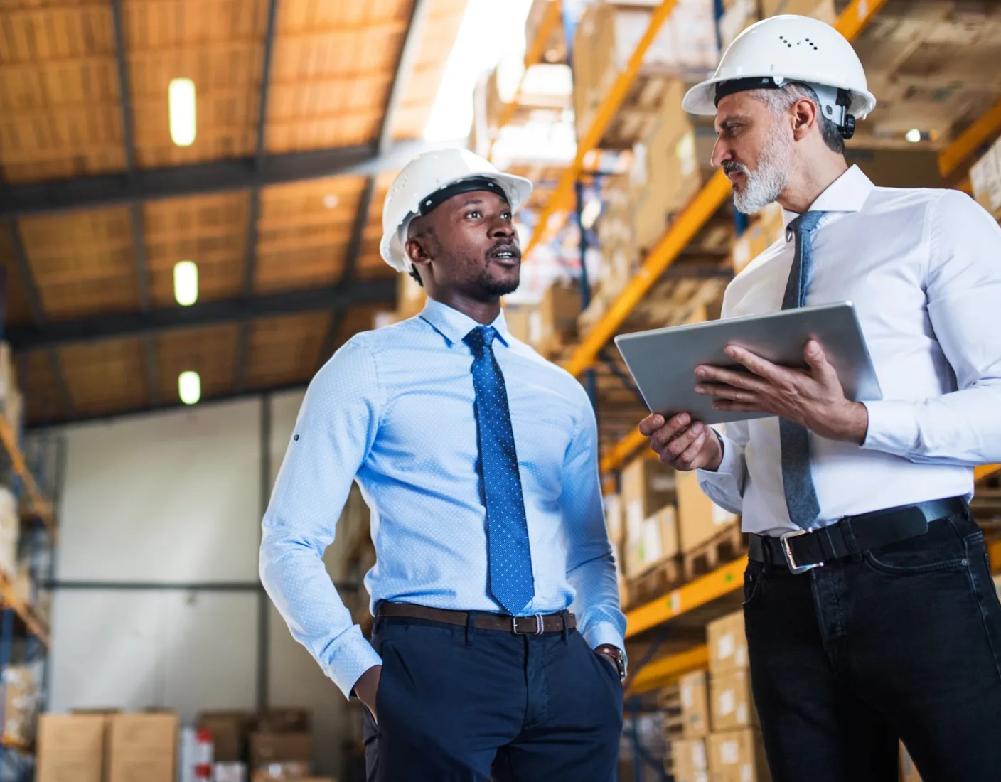 Two men wearing hard hats and ties discuss something in a large warehouse. One man holds a tablet. Shelves with boxes are in the background.