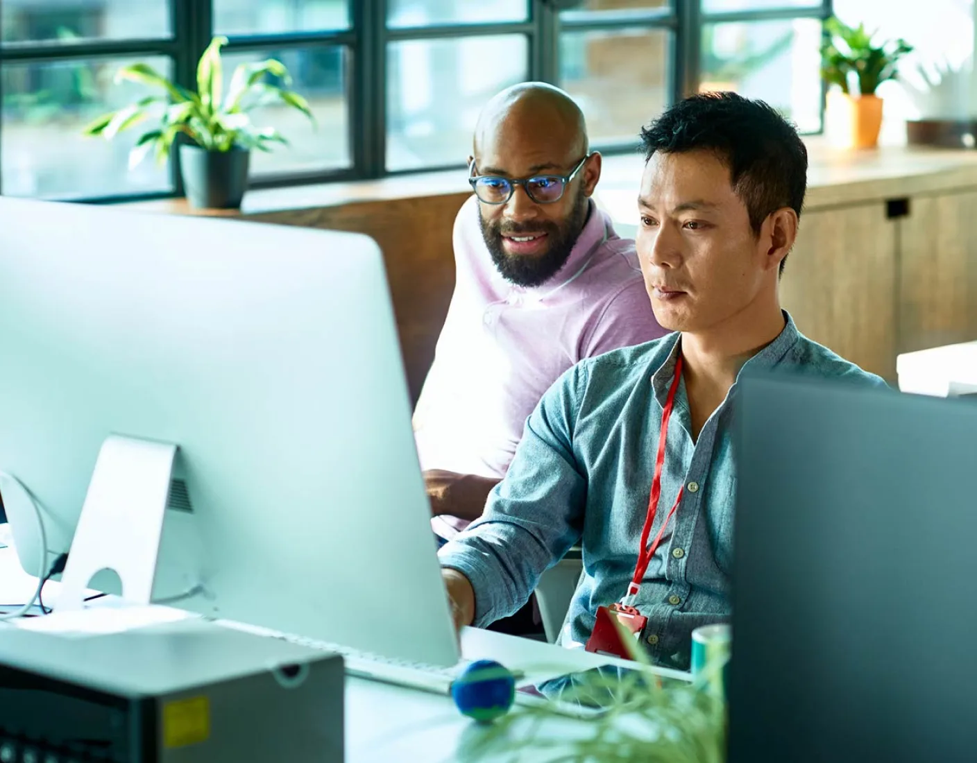 Two men are working at a desk with a computer screen in an office setting. One man is sitting and focused on the screen, while the other is standing and observing. Plant pots are visible in the background.