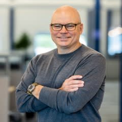 Headshot of blog author Chris Powell. He wears glasses and a gray sweater and stands with his arms crossed, smiling, in a modern indoor setting with blurred office background.