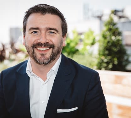 Headshot of blog author James Fisher. He is bearded and in a navy blue suit and white shirt. He smiles while posing outdoors with greenery and buildings in the background.