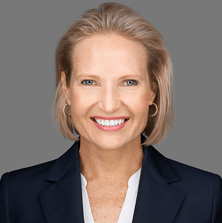 Headshot of blog author Julie Kae. She has short blonde hair and blue eyes and smiles at the camera, wearing a black top and a necklace, with a plain light-colored background.